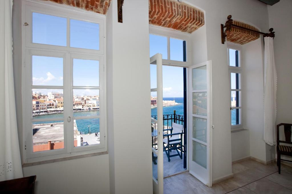  Interior view of a bright Mediterranean room with white walls and exposed brick archways. Large French windows and a glass door open to a small balcony overlooking the Venetian harbor of Chania, Crete. Through the windows, you can see the turquoise sea, colorful waterfront buildings, and the historic lighthouse in the distance. The room features traditional architectural elements like exposed brick lintels and white-painted woodwork.