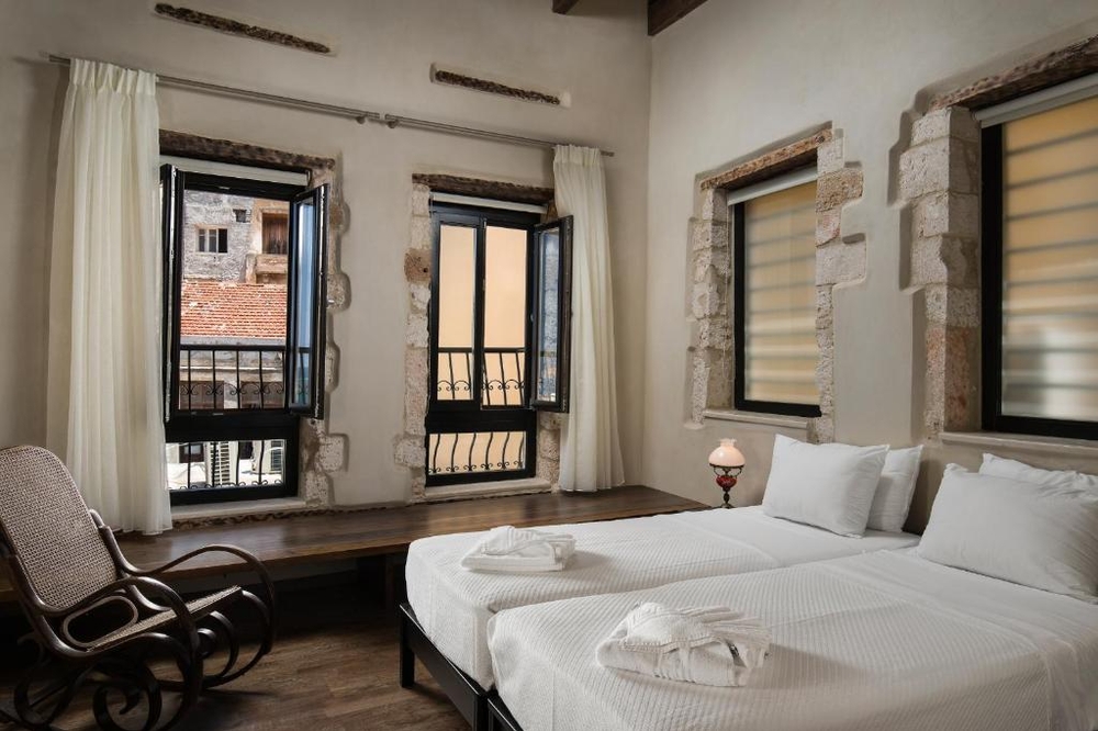 A rustic-elegant hotel room with exposed stone window frames and wooden ceiling beams. The room features twin beds with crisp white linens and bathrobes, dark wood floors, and floor-to-ceiling French windows with black frames and decorative iron balconies. White curtains frame the windows, and a vintage-style rocking chair sits by the window bench. The room combines traditional Mediterranean architectural elements with modern comfort.