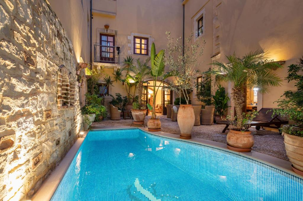 A charming courtyard pool in a traditional Mediterranean building, illuminated at dusk. The small mosaic-tiled pool is surrounded by stone walls and cream-colored buildings with wooden-framed windows and balconies. Large terracotta pots containing palms, banana plants, and olive trees line the poolside, creating a lush garden atmosphere. Uplighting on the walls and warm interior lights from the windows create an intimate evening ambiance.