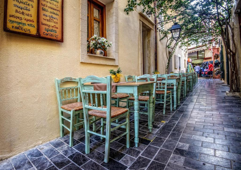 A charming narrow alley in Chania's old town with traditional taverna seating arranged along a cream-colored wall. The chairs are painted in a distinctive Mediterranean mint green with woven rush seats, paired with matching tables. A menu board hangs on the wall, and a flower pot sits in a window niche. At the end of the alley, there's a glimpse of a tourist shop with clothing displays. The stone-paved street and overall atmosphere captures the quintessential Greek taverna setting.