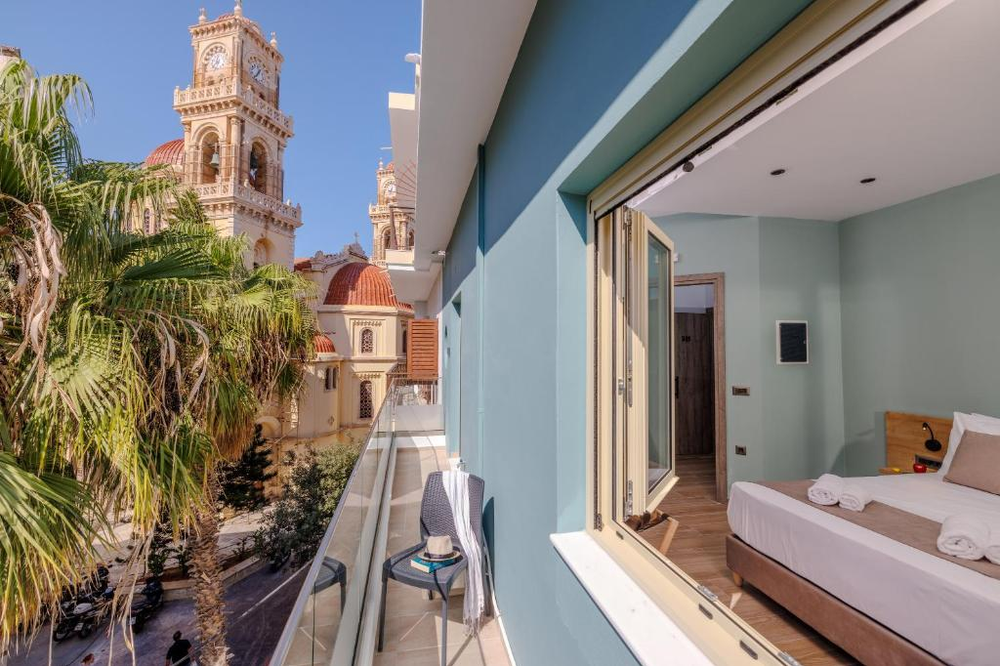 A hotel room with a balcony in Heraklion, Crete, offering a stunning view of the Agios Minas Cathedral. The room features mint-green walls, hardwood floors, and a modern bed with white linens. Through the sliding glass doors, the balcony offers a close-up view of the cathedral's ornate bell towers and copper-colored domes. A palm tree frames the historic church architecture, creating a perfect blend of Mediterranean and Byzantine aesthetics.