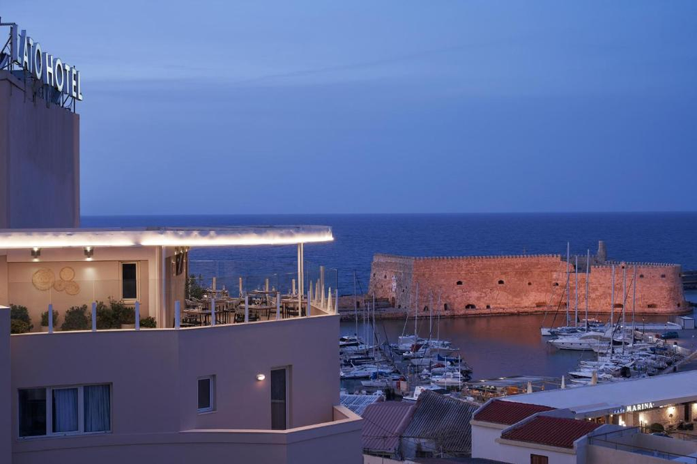  Evening view of Heraklion harbor featuring the historic Koules Fortress (Rocca al Mare) illuminated in warm light. The Venetian fortress appears reddish-orange against the deep blue twilight sky and sea. In the foreground is part of a modern hotel building with lit terraces, and the harbor is filled with moored sailboats and yachts. The image captures the blend of historic architecture and modern marine life characteristic of Crete's main port city.