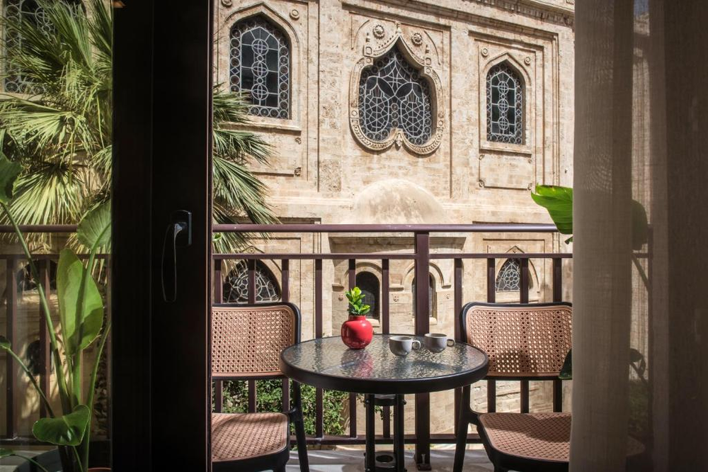 A charming hotel balcony view in Heraklion featuring ornate Gothic-style church windows with decorative stonework across the street. The balcony is furnished with a small round table and two wicker chairs, with coffee cups and a red potted plant on the table. Palm fronds and tropical plants frame the view, creating an intimate setting facing the historic church architecture. CopyRetry