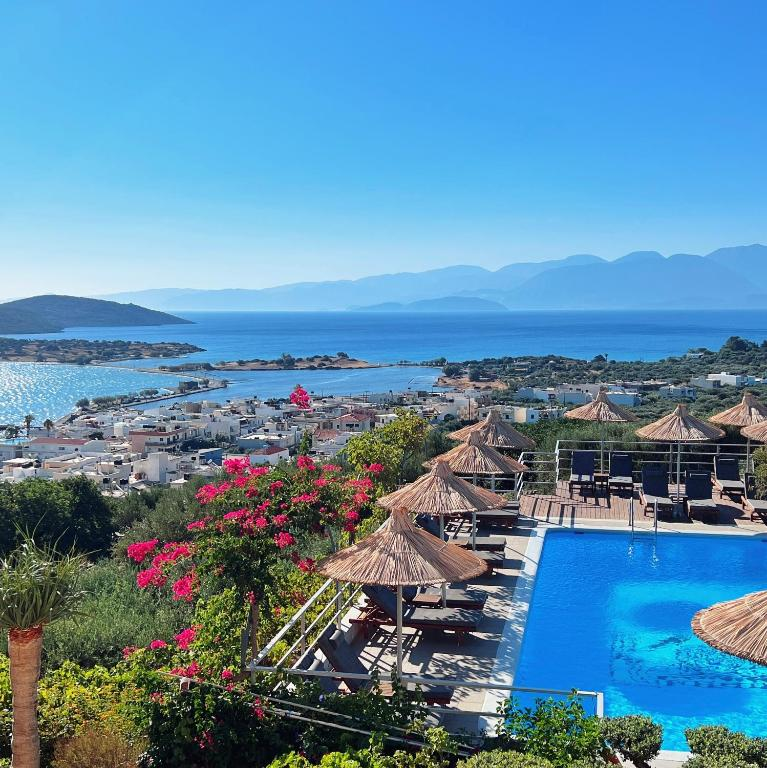 An elevated view of a coastal town in Crete from a hotel pool terrace. In the foreground is a bright blue swimming pool lined with thatched umbrellas and sun loungers. Vibrant pink bougainvillea flowers frame the view of the white buildings below, which stretch down to a Mediterranean bay. Mountains rise in the background across the azure water, and the clear blue sky completes this quintessential Greek island scene.