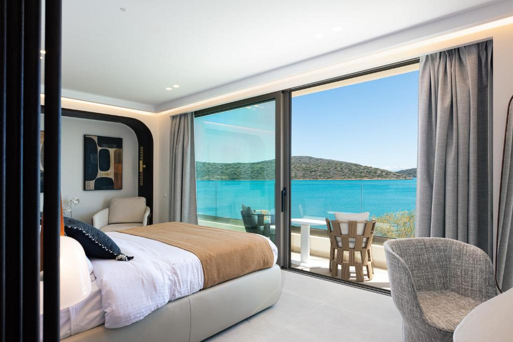 A luxurious modern hotel room featuring floor-to-ceiling sliding glass doors that frame a stunning view of turquoise Mediterranean waters and green hills. The room is decorated in a contemporary style with a white and beige color scheme, including a platform bed with white linens and a tan throw, grey upholstered chairs, and modern artwork. A private balcony with glass railing and casual seating extends the living space outdoors. The space combines sleek design with panoramic sea views.