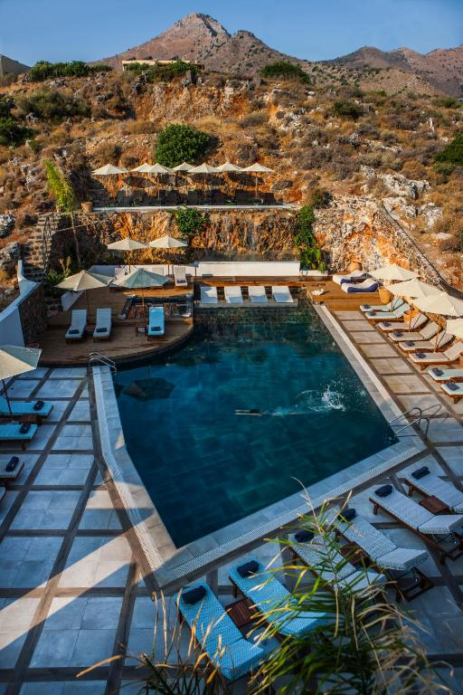 A luxury hotel pool area built into a rocky hillside in Crete. The L-shaped pool is surrounded by white sun loungers and cream-colored umbrellas on multiple terraced levels. The rugged mountain landscape rises dramatically behind the pool, with its golden-brown cliffs contrasting against the deep blue sky. The design seamlessly integrates modern amenities with the natural terrain, with someone visible swimming in the deep blue water.