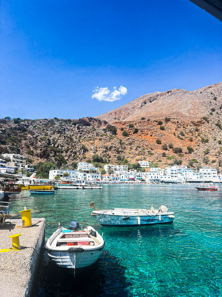 The picturesque village of Loutro in Crete, accessible only by boat, nestled at the foot of rugged mountains. Small fishing boats bob in the crystal-clear turquoise waters of the harbor. The village's characteristic white-and-blue buildings climb up the rocky hillside in tiers. Under a bright blue sky with a single white cloud, the scene captures the remote, tranquil nature of this car-free coastal settlement.