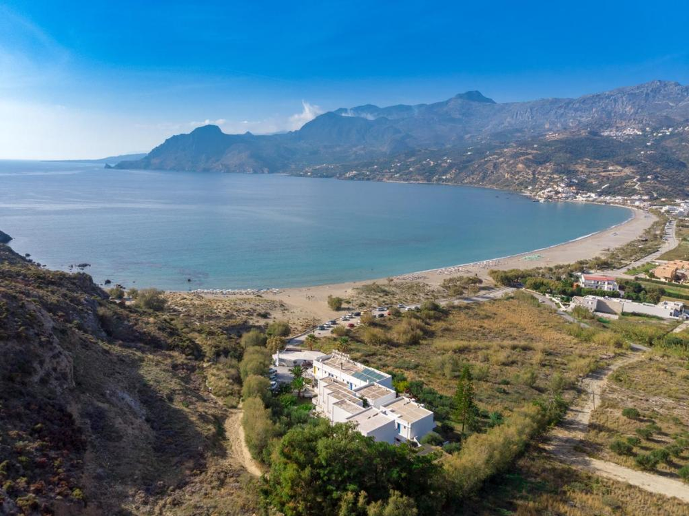 An aerial view of Plakias Bay in southern Crete, showing a curved beach stretching along a dramatic coastline with mountains rising steeply from the sea. In the foreground, a white modern hotel complex sits nestled in greenery, while the expansive bay curves around with turquoise waters meeting the sandy beach. The rugged mountains create a dramatic backdrop against the blue sky, and scattered development can be seen along the coastline.