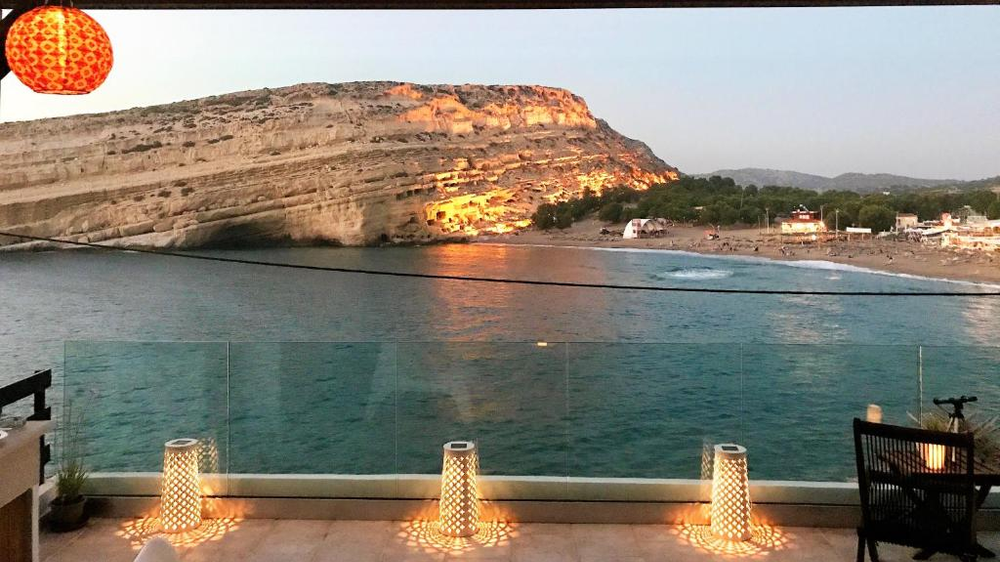 Evening view from a hotel terrace in Matala, Crete, overlooking the famous beach and cliffs. The limestone cliffs, known for their ancient caves, are illuminated by the setting sun, giving them a golden glow. The terrace features a glass balustrade and decorative lanterns casting patterns on the floor. A red Chinese lantern appears in the top corner, and the turquoise waters of the bay reflect the evening light.