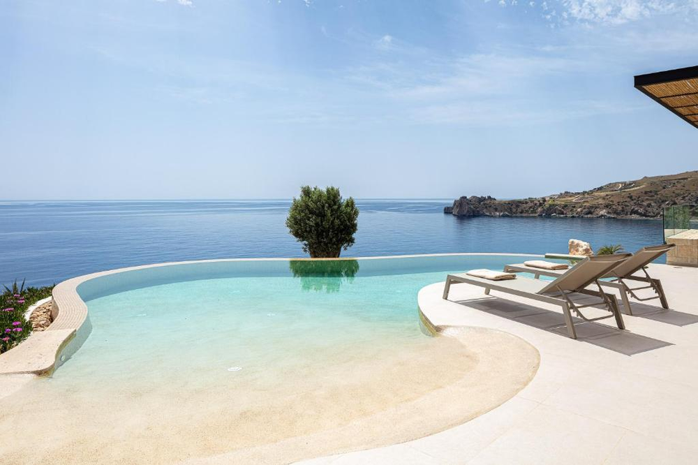 A luxurious infinity pool overlooking the Mediterranean Sea in Crete. The pool has a unique beach-entry design with a gradual slope and curved edges, creating the illusion of merging with the sea horizon. A single olive tree stands in the middle of the pool, and modern sun loungers are placed on the white terrace. The view extends to rocky coastal cliffs in the distance under a clear blue sky.