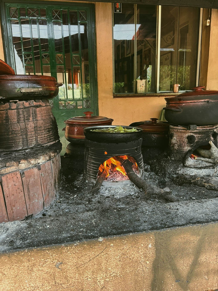 Traditional Cretan cooking setup at Dounias Taverna, showing several clay pots and cooking vessels arranged on brick and stone hearths. A live fire can be seen burning beneath one of the pots, with flames visible through the opening. The setup demonstrates authentic slow-cooking methods typical of traditional Greek cuisine. The cookware appears well-used and seasoned, suggesting regular use for preparing traditional dishes.