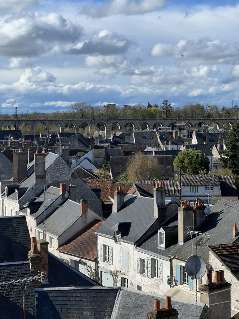 A scenic view of Beaugency, France, featuring a charming cluster of slate-roofed houses with brick chimneys in the foreground. In the background, an elegant stone viaduct arches over the landscape, set against a sky filled with soft clouds.