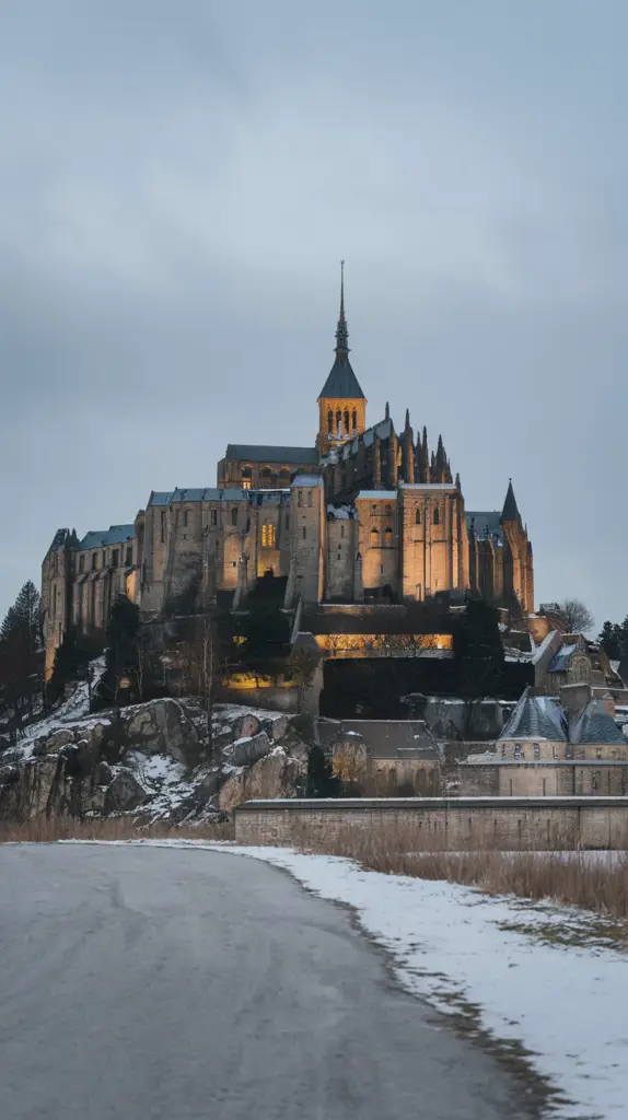 This image shows a majestic hilltop castle or monastery illuminated warmly against a cold wintery backdrop. The building features intricate medieval architecture with a central spire rising into the cloudy sky. The foreground includes a snowy path and barren landscape, enhancing the serene and slightly