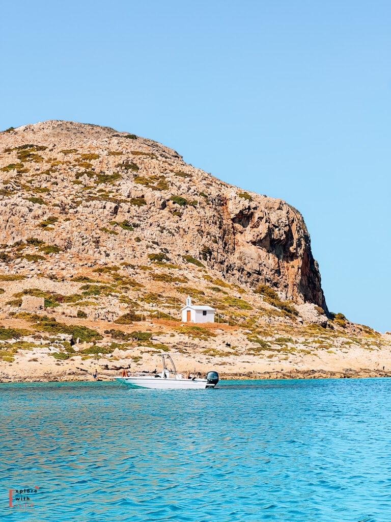 A view along the Cretan coastline showing a small white chapel perched on a rocky cliff face. In the foreground, a white speedboat floats on brilliant turquoise Mediterranean waters. The limestone cliff rises dramatically against a clear blue sky, with sparse vegetation dotting its rocky surface. This scene was captured en route to Balos Beach, showcasing the characteristic landscape of the Greek islands.