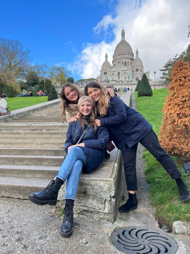 Three young women pose cheerfully on the steps leading up to the Sacré-Cœur Basilica in Paris. They are dressed casually in winter wear - one in a cream sweater, and two in navy blue jackets with jeans and boots. The iconic white domed church stands majestically in the background against a blue sky with white clouds. Manicured topiary bushes line the grassy slopes on either side of the steps. The image captures a warm family moment with three sisters smiling together at this famous Montmartre landmark.