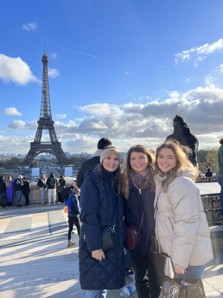 Three young women pose together at the Trocadero platform with the Eiffel Tower in the background on a sunny winter day. They're dressed warmly - one in a long navy quilted coat and beanie, one in dark winter wear with a scarf, and one in a cream-colored puffer jacket. The iconic tower stands against a bright blue sky with white clouds, and other tourists can be seen in the background of this popular viewpoint.