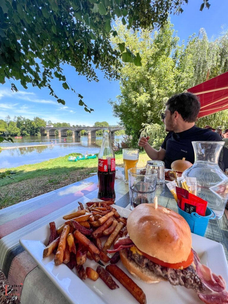 A riverside lunch scene at La Guinguette du Pont de Cénac, with a juicy burger and french fries in the foreground on a white plate. Behind the meal, the historic Cénac bridge spans the Dordogne River where colorful kayaks can be seen on the riverbank. A Coca-Cola bottle, beer glass, and water carafe sit on the outdoor table, while tree leaves frame the sunny summer scene from above. A diner in a navy shirt enjoys the casual riverside dining experience.