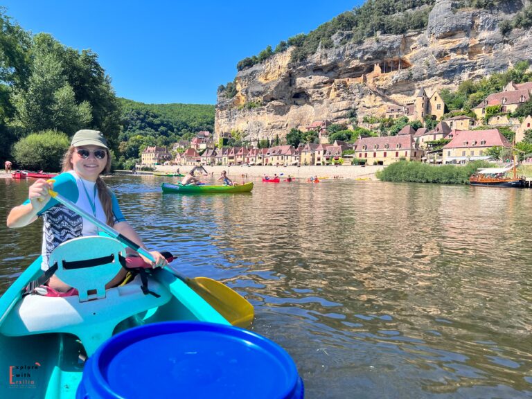 A woman paddles a turquoise canoe on the Dordogne River, smiling at the camera while wearing a sun hat and sunglasses. The dramatic backdrop shows La Roque-Gageac, one of France's most beautiful villages, with its honey-colored buildings built into the towering limestone cliff. Other canoeists can be seen on the river, and a traditional gabarre boat is moored along the village waterfront. The scene captures the bustling summer activity on this scenic stretch of the Dordogne.