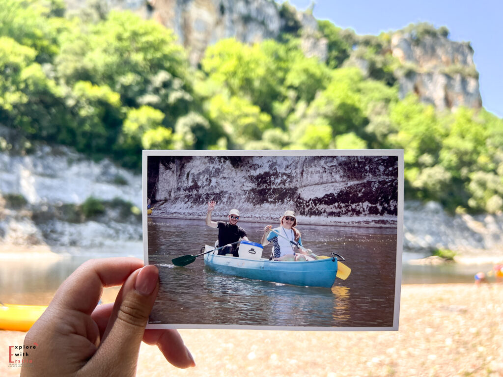 A hand holds up a printed photograph showing a couple in a turquoise canoe on the Dordogne River, both smiling and waving at the camera. The photo is being held up against the actual river scene, creating a "photo within a photo" effect. The limestone cliffs and green foliage visible in both the printed photo and the background create a matching perspective that connects past and present moments of the river journey.