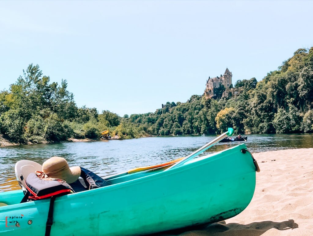 A turquoise canoe rests on a sandy beach along the Dordogne River, with a sun hat visible inside. In the background, the medieval Château de Montfort perches dramatically on a forested cliff overlooking the river. The scene captures a peaceful summer day with clear blue skies and lush green trees lining the riverbank. Another canoe can be seen on the calm waters in the distance.