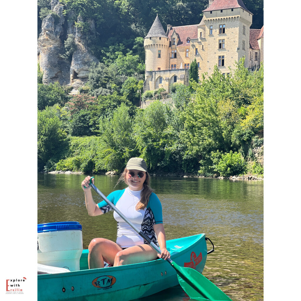 Ersilia paddling a turquoise canoe on the Dordogne River, smiling at the camera while wearing a sun hat and sunglasses. Behind her, the Château de la Malartrie rises dramatically from the limestone cliffs, its honey-colored stone towers and turrets standing out against the lush green foliage. The castle's medieval architecture and its dramatic setting above the river create a quintessential Dordogne Valley scene.