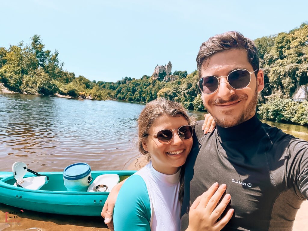 A couple takes a selfie while canoeing on the Dordogne River, with Château de Montfort visible on the clifftop in the background. They're both wearing sunglasses and smiling at the camera, their turquoise canoe visible in the frame. The day appears sunny and bright, with the castle and forested hills creating a picturesque backdrop for their river adventure.