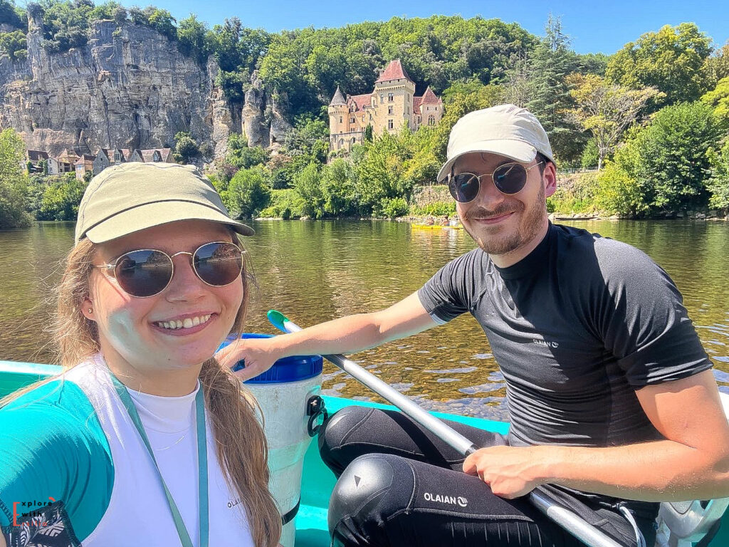 A couple takes a selfie from their turquoise canoe on the Dordogne River, with the village of La Roque-Gageac in the background. Both are wearing baseball caps and sunglasses, smiling at the camera. The village's distinctive honey-colored buildings are built into the limestone cliff face, with the château rising above. The scene captures a sunny day on the river with clear reflections in the calm water.