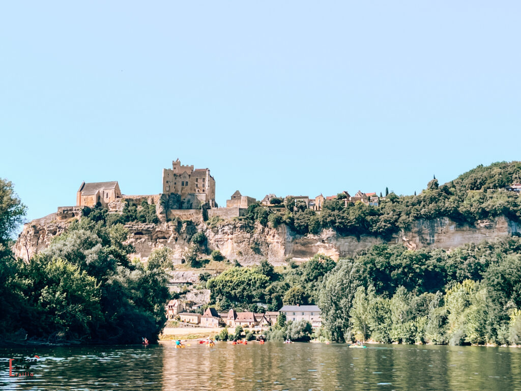 View of Château de Beynac from the Dordogne River, with the medieval fortress dramatically positioned atop limestone cliffs. The castle and its chapel stand prominently against a bright blue sky, with the village houses terraced down the hillside below. Colorful kayaks dot the river in the foreground, where tourists enjoy water activities on a sunny day. The scene illustrates the perfect blend of historic architecture, natural landscape, and modern tourism that characterizes the Dordogne Valley.