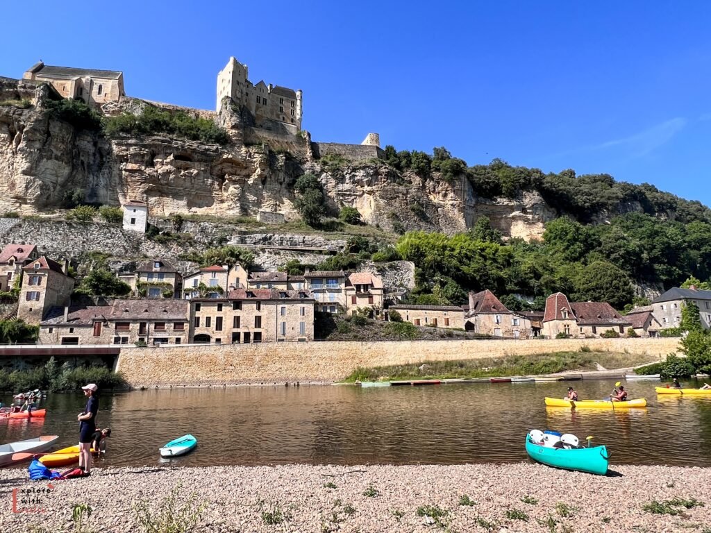  riverside view of the village of Beynac-et-Cazenac in the Dordogne Valley. The medieval Château de Beynac crowns the limestone cliff, with the village's stone houses terraced below it. The riverbank is busy with kayakers and canoeists, with colorful kayaks dotting both the pebbly beach and the water. The traditional honey-colored stone architecture stands in dramatic contrast against the bright blue sky, while a stone retaining wall separates the village from the river.