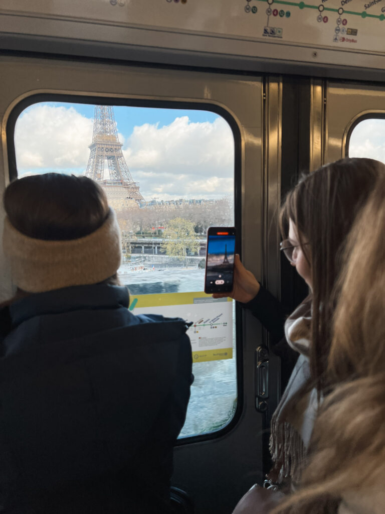 View from inside a Paris Metro, where two passengers are looking out the window at the Eiffel Tower visible in the distance. One passenger is taking a photo of the tower with their phone. The window frames the Eiffel Tower against a cloudy sky, with the Seine River visible below. A transit map is partially visible at the top of the train car.