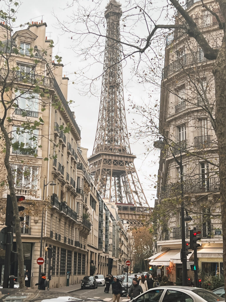 A charming Parisian street view with the Eiffel Tower rising between classic Haussmann-style apartment buildings. The scene is captured during autumn or winter, with bare tree branches framing the view. The cream-colored buildings feature traditional French balconies and architectural details. At street level, there are parked cars, traffic lights, and pedestrians walking along the sidewalk. The overall image has a soft, muted color palette typical of Paris.