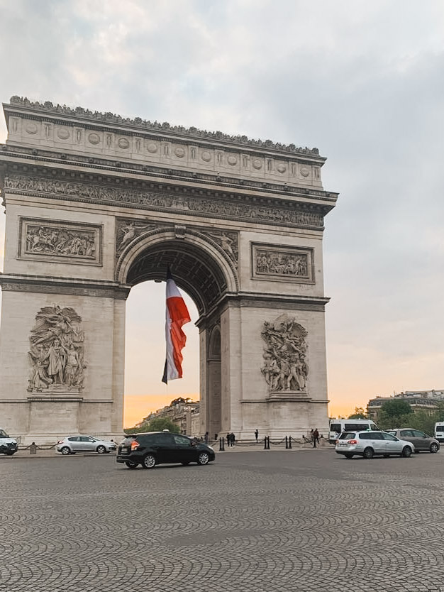 The Arc de Triomphe in Paris at sunset, with a large French tricolor flag flying through its central arch. The monument's intricate relief sculptures and architectural details are visible on its limestone facade. Cars circle around the cobblestoned Place Charles de Gaulle, and the sky shows warm sunset colors behind the arch. The scene captures both the monument's imposing scale and its role as a symbol of French patriotism.