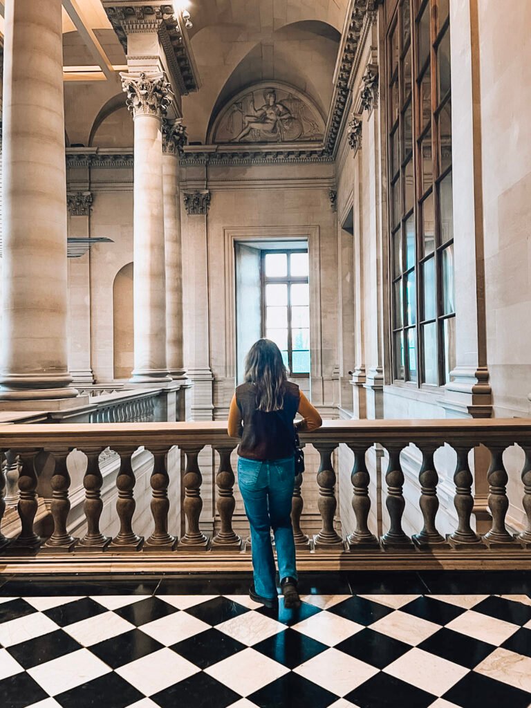 Interior view of the Louvre Museum, showing a person standing at an ornate balustrade looking out into a grand hallway. The space features classical architecture with tall columns, decorative capitals, and arched ceilings with relief sculptures. The floor has a distinctive black and white checkered pattern. The person is wearing blue jeans and a dark top, photographed from behind as they take in the majestic corridor with its high windows and architectural details.