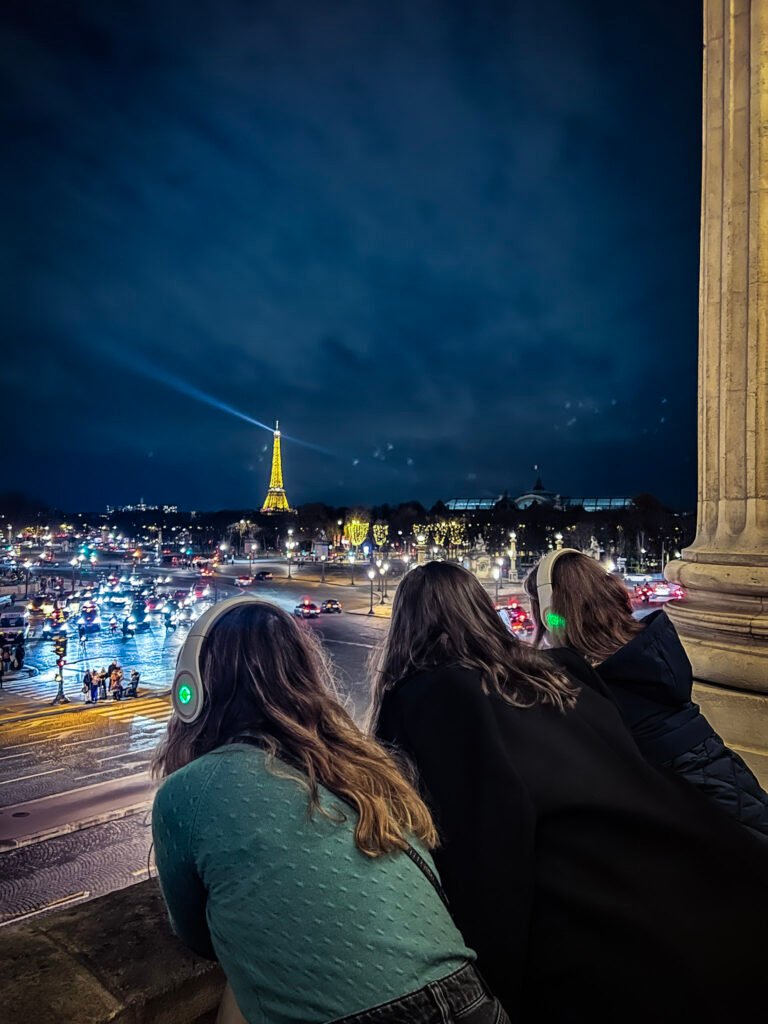 Nighttime view from the Hotel de la Marine balcony in Paris, showing three people wearing illuminated headphones while looking out at the illuminated Eiffel Tower in the distance. The scene captures the bustling Place de la Concorde below with streams of car headlights and taillights creating light trails. The Eiffel Tower glows golden against the dark blue night sky and appears to be displaying its signature sparkle light show. The viewers are photographed from behind as they lean against the historic building's balcony to take in the iconic Parisian nightscape.