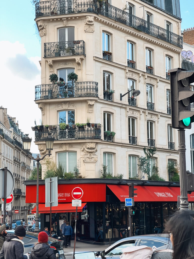 A classic Parisian corner building with typical Haussmann architecture, featuring cream-colored stone and ornate wrought iron balconies on each floor. The building has decorative moldings and potted plants on its balconies. At street level, there's a business with distinctive red awnings. Street signs, lampposts, and pedestrians are visible at ground level, creating a quintessential Parisian street scene.