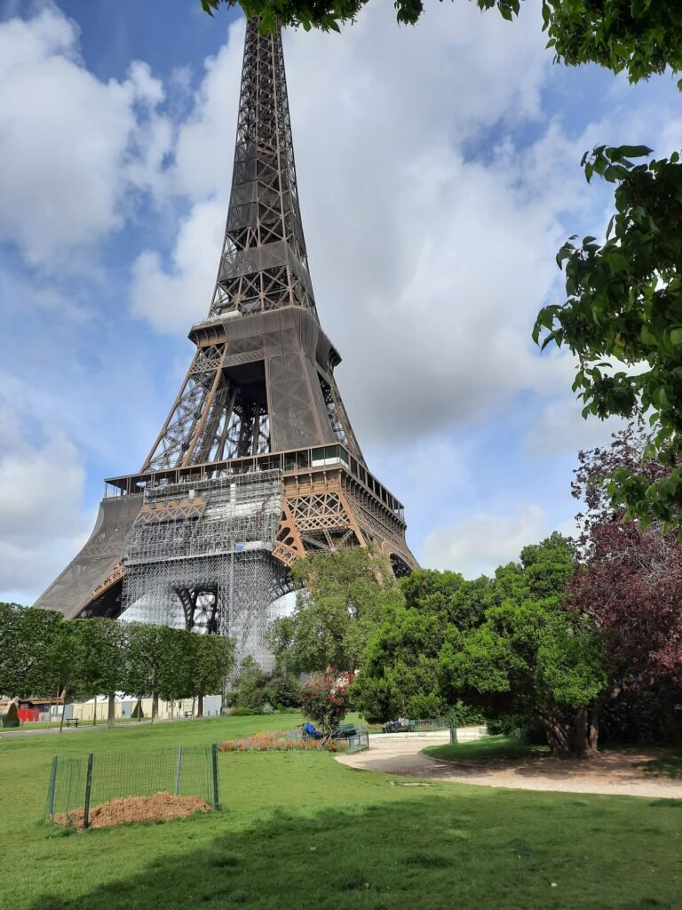 A close-up view of the Eiffel Tower's base and lower section, showing ongoing renovation work with white scaffolding on one side. The tower is photographed from the Champ de Mars garden, which features green lawns, trees, and some construction fencing around a dirt area. The sky is bright blue with white clouds, and the tower's iron lattice work is clearly visible against it. Some flowering plants and shrubs can be seen in the landscaped gardens beneath the tower.