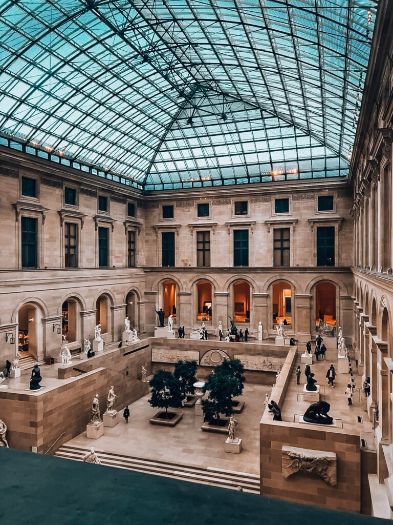 Interior courtyard of the Louvre Museum featuring a glass pyramid ceiling and neoclassical architecture. The space displays numerous classical sculptures, both white marble and dark bronze, arranged along tiered levels. Small ornamental trees in planters sit in the center of the courtyard. The stone walls feature arched galleries and decorative elements, with warm lighting visible through the archways. Visitors can be seen walking through the space, appearing small in scale compared to the grand architecture. The scene is captured from an elevated viewpoint, showing the symmetrical layout of this historic gallery space.