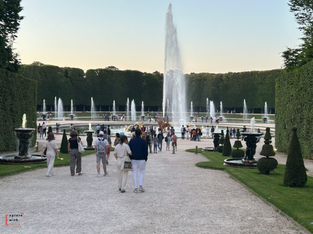 A bustling crowd at the Gardens of Versailles during the summer night fountain shows, with the illuminated Palace of Versailles visible in the distance. Visitors stroll through tree-lined paths and gather near fountains under a clear evening sky, enjoying the lively atmosphere of this iconic French landmark.