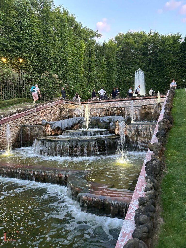 A tiered fountain in the Gardens of Versailles, captured during the summer night fountain shows. Water cascades down stone basins, surrounded by lush greenery and illuminated by soft lights. Visitors are seen walking along the pathways, adding a lively yet serene ambiance to the historic setting.