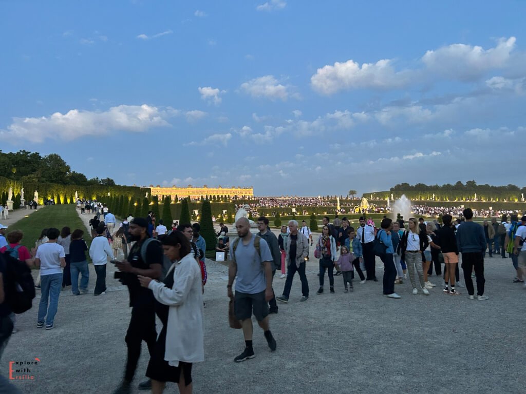 A bustling crowd at the Gardens of Versailles during the summer night fountain shows, with the illuminated Palace of Versailles visible in the distance. Visitors stroll through tree-lined paths and gather near fountains under a clear evening sky, enjoying the lively atmosphere of this iconic French landmark.