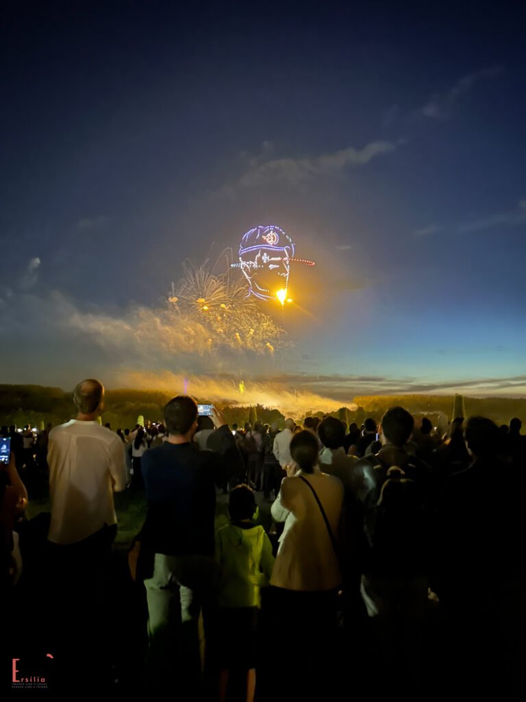 A nighttime fireworks display at Versailles featuring a large illuminated design in the sky that appears to be a pilot's face with a cap, created with drone lights or light projections. Below, a crowd of spectators watches as golden fireworks explode against the dark sky. The image captures a special celebration for the French Air Force's 90th anniversary, with people taking photos and children on shoulders to view the spectacle.