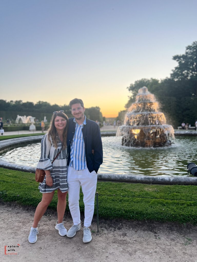 A couple smiling in front of a lit fountain at the Gardens of Versailles during a summer evening, part of the night fountain shows. The man wears white pants with a striped shirt and dark jacket, and the woman is in a patterned dress with a small woven bag. The golden sunset and lush gardens create a serene, elegant backdrop.