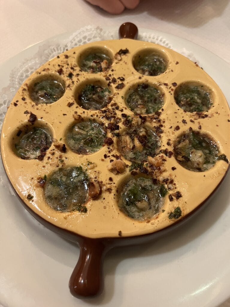  A traditional French escargot dish served in a yellow ceramic escargot plate with twelve indentations. The snails are prepared in a classic style with garlic, parsley, and butter, garnished with toasted breadcrumbs scattered across the plate. The specialized serving dish has a brown handle and sits on a white plate with a decorative lace doily underneath.