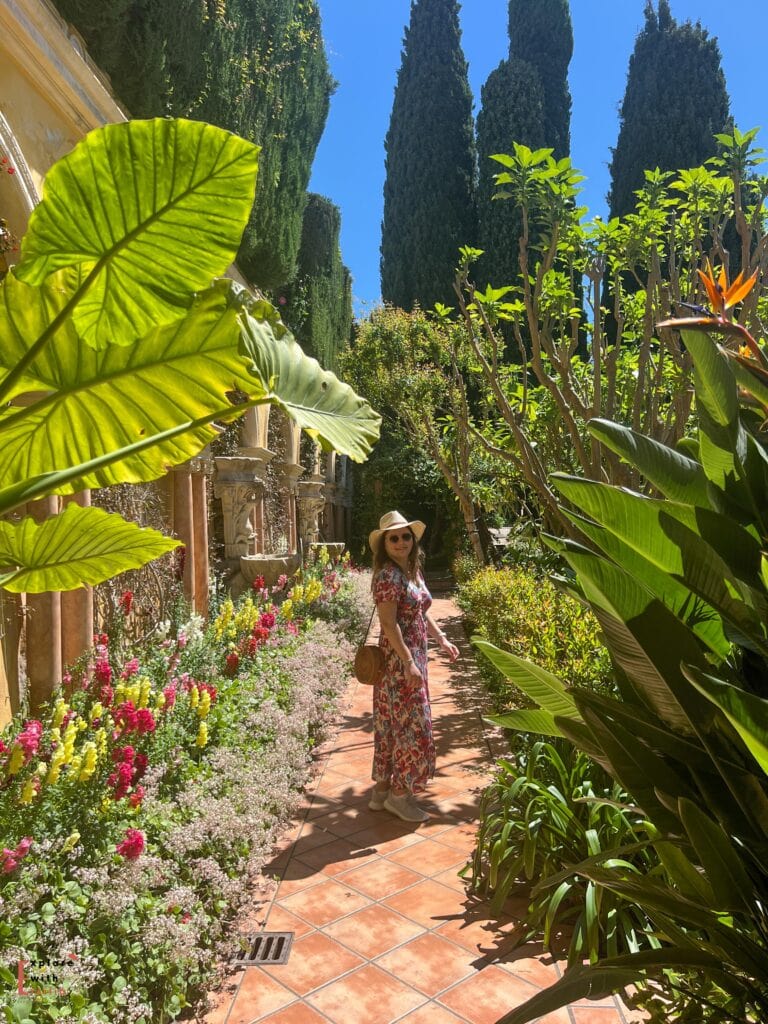 Woman in a floral dress and straw hat standing in Villa Ephrussi de Rothschild's Spanish garden, with tall cypress trees, large tropical leaves in the foreground, and colorful flower beds along the terracotta-tiled path. The scene captures the villa's diverse botanical collection under bright Mediterranean sunlight