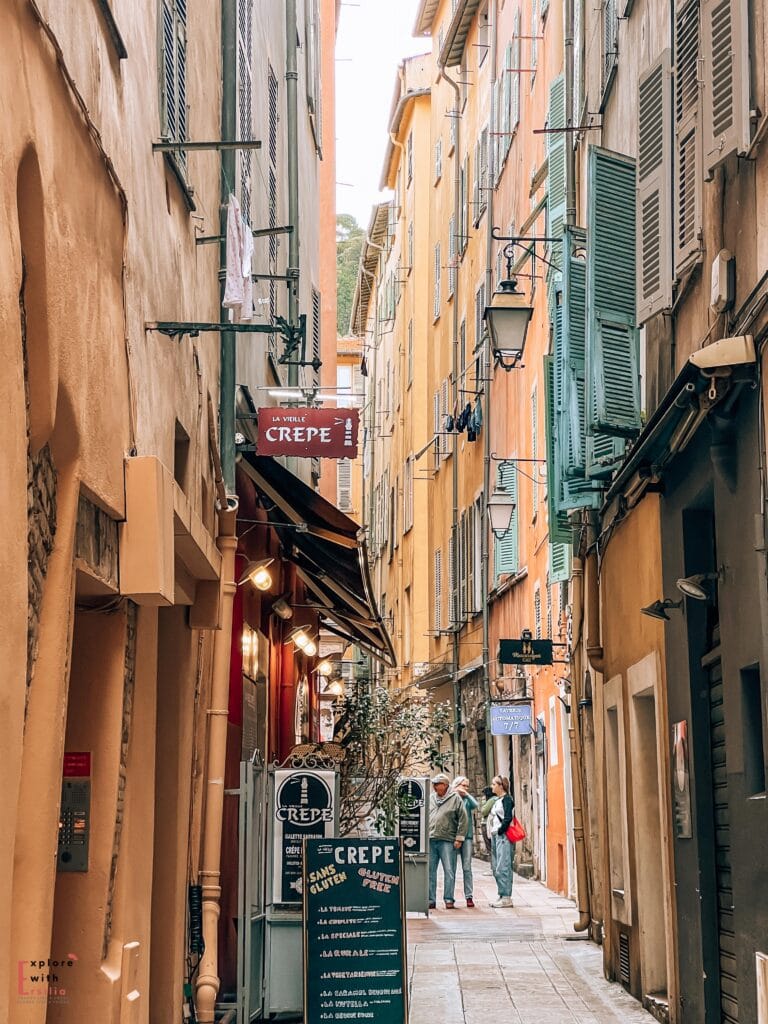 A narrow cobblestone alley in Nice's old town, with 'La Vieille Crepe' restaurant sign hanging from a rustic bracket. The atmospheric street is lined with tall buildings in warm peach and orange tones with traditional blue-green shutters. String lights hang overhead, and a chalkboard menu advertising gluten-free crepes stands outside the restaurant. A few pedestrians chat in the distance.