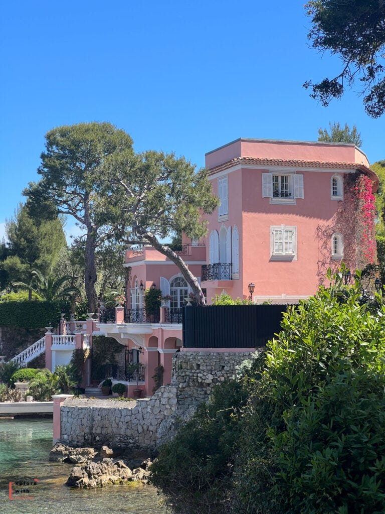 A pink villa perched on Saint-Jean-Cap-Ferrat's waterfront, featuring Mediterranean architecture with terracotta roof tiles, white balustrades, and arched details. The multi-level building is surrounded by pine trees and landscaped gardens, with stone walls descending to the water's edge.