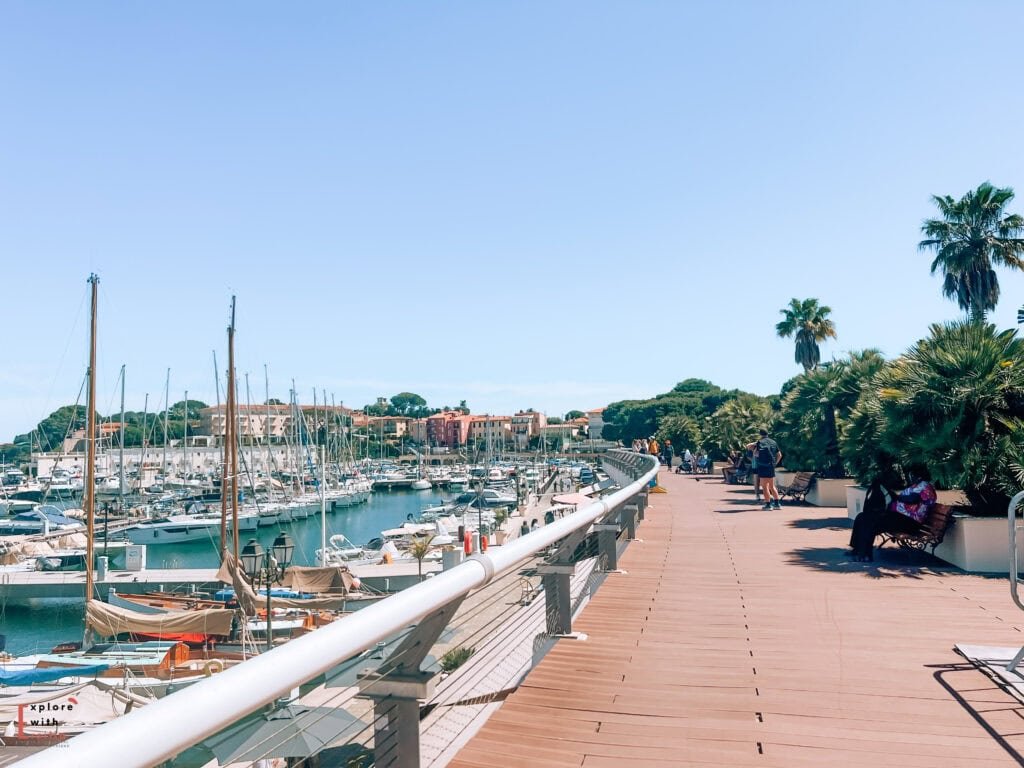 View of Saint-Jean-Cap-Ferrat's marina boardwalk lined with boats and yachts. The wooden walkway features white railings, with colorful Mediterranean buildings visible in the background under a bright blue sky