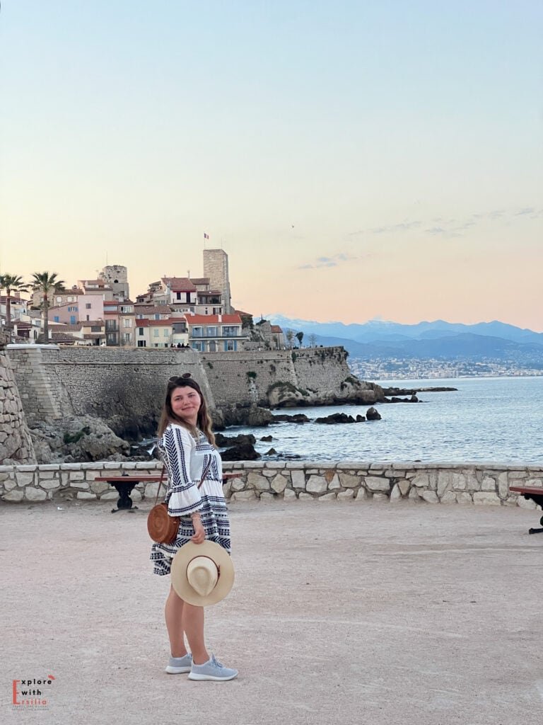 Sunset view of Antibes' medieval ramparts and old town, with a woman in a patterned black and white dress holding a straw hat. The historic stone fortifications and pastel-colored buildings are backdrop to the Mediterranean Sea and Alpine foothills in the distance, captured during golden hour.