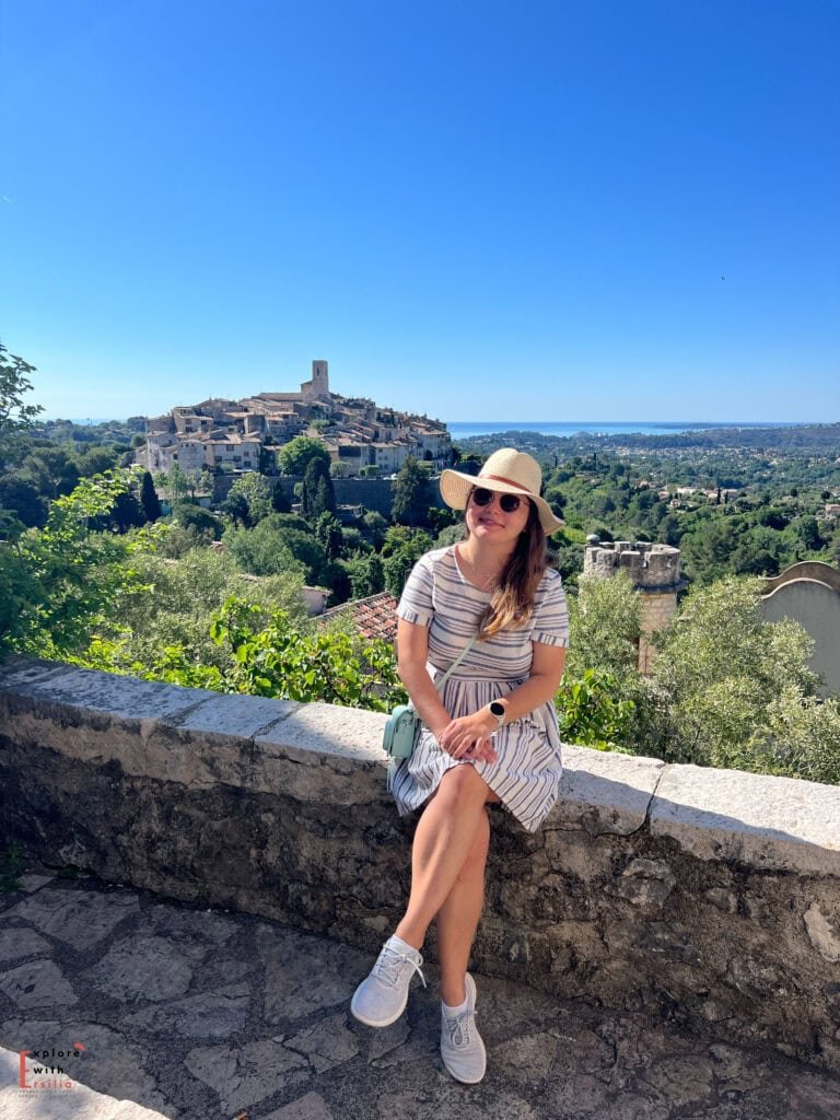 Portrait of a woman in a striped dress and straw hat sitting on a stone wall, with the medieval hilltop village of Saint-Paul-de-Vence in the background. The historic village's church tower rises above clustered stone buildings, while the Mediterranean coast and surrounding hills stretch to the horizon on a clear, sunny day.