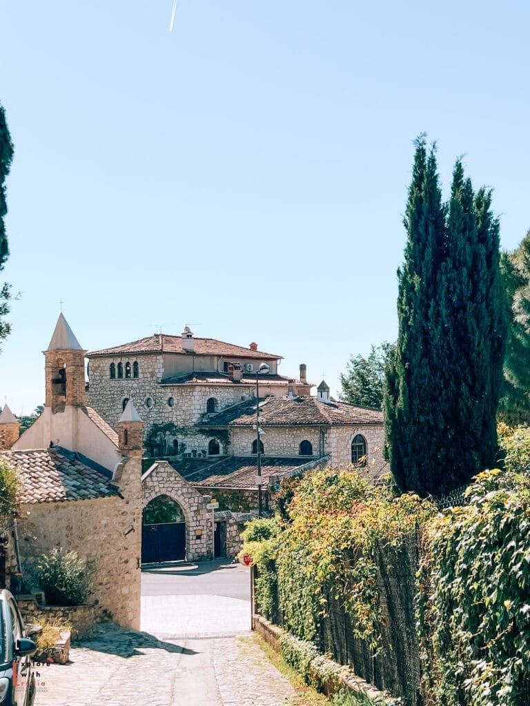 The Colombe d'Or hotel and restaurant in Saint-Paul-de-Vence, known for its art collection and famous past guests. The stone building features rustic architecture with terracotta roof tiles, arched entrances, and tall cypress trees framing the scene against a bright blue sky.